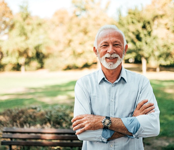 An older man standing outside with his arms crossed in front of him smiling and enjoying his new teeth